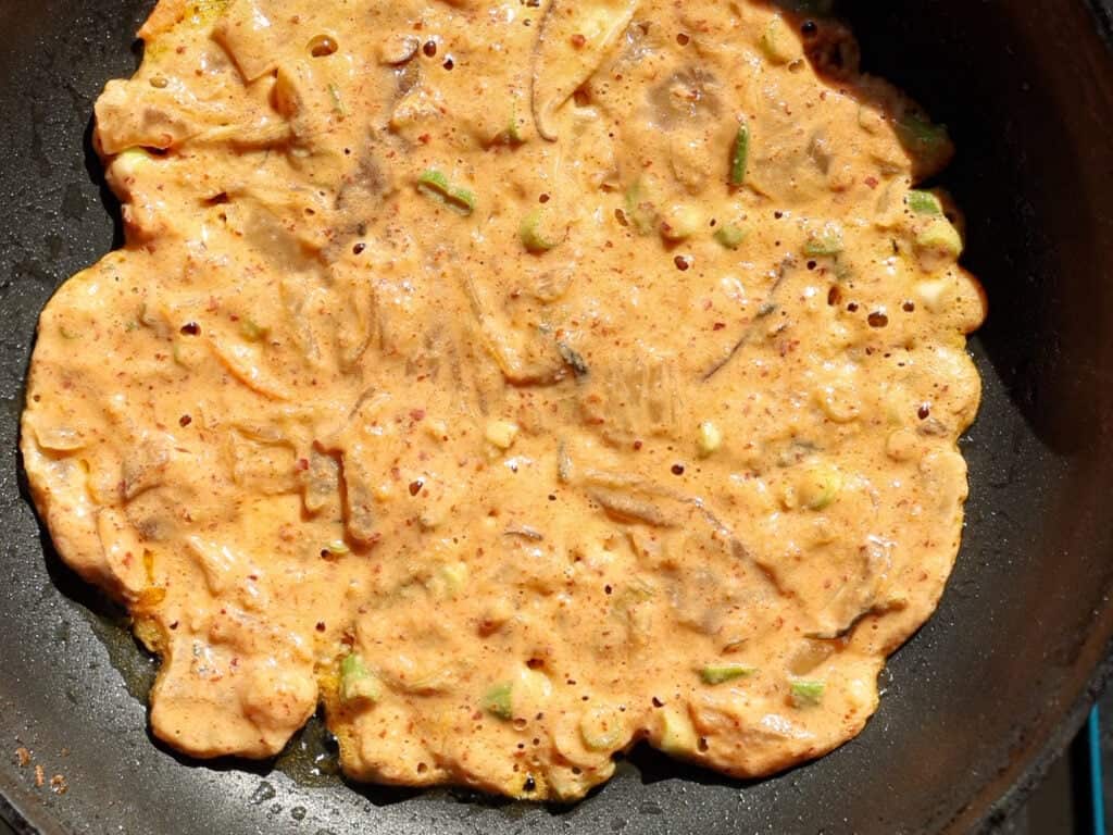 A close-up of a Korean pancake (jeon) cooking in a black pan, with visible bits of vegetables in the orange batter. The pancake is partially cooked, with some bubbles and uneven edges.